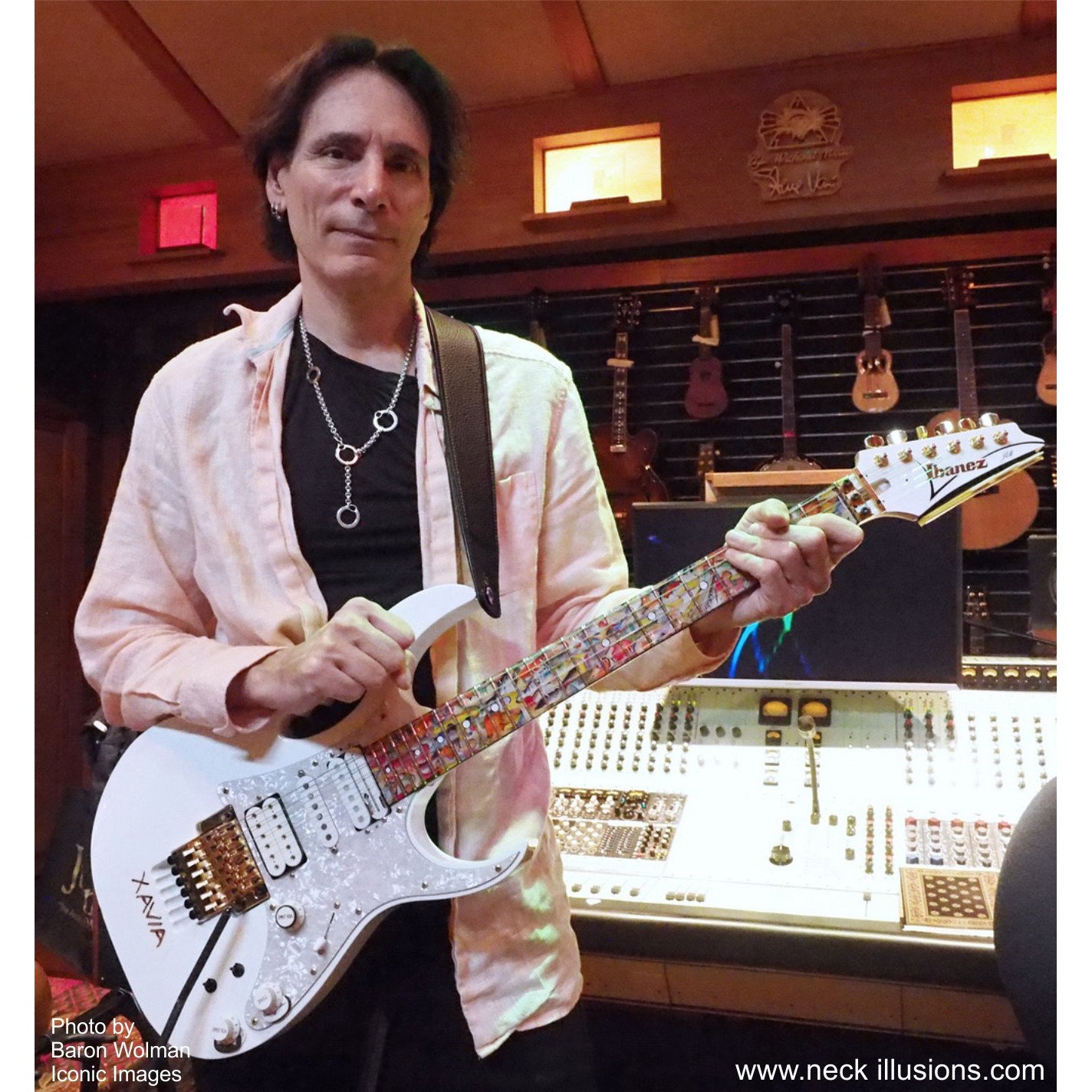 Image of steve vai in the recording studio holding a white electric guitar with a colorful fret protector on the neck of the guitar.