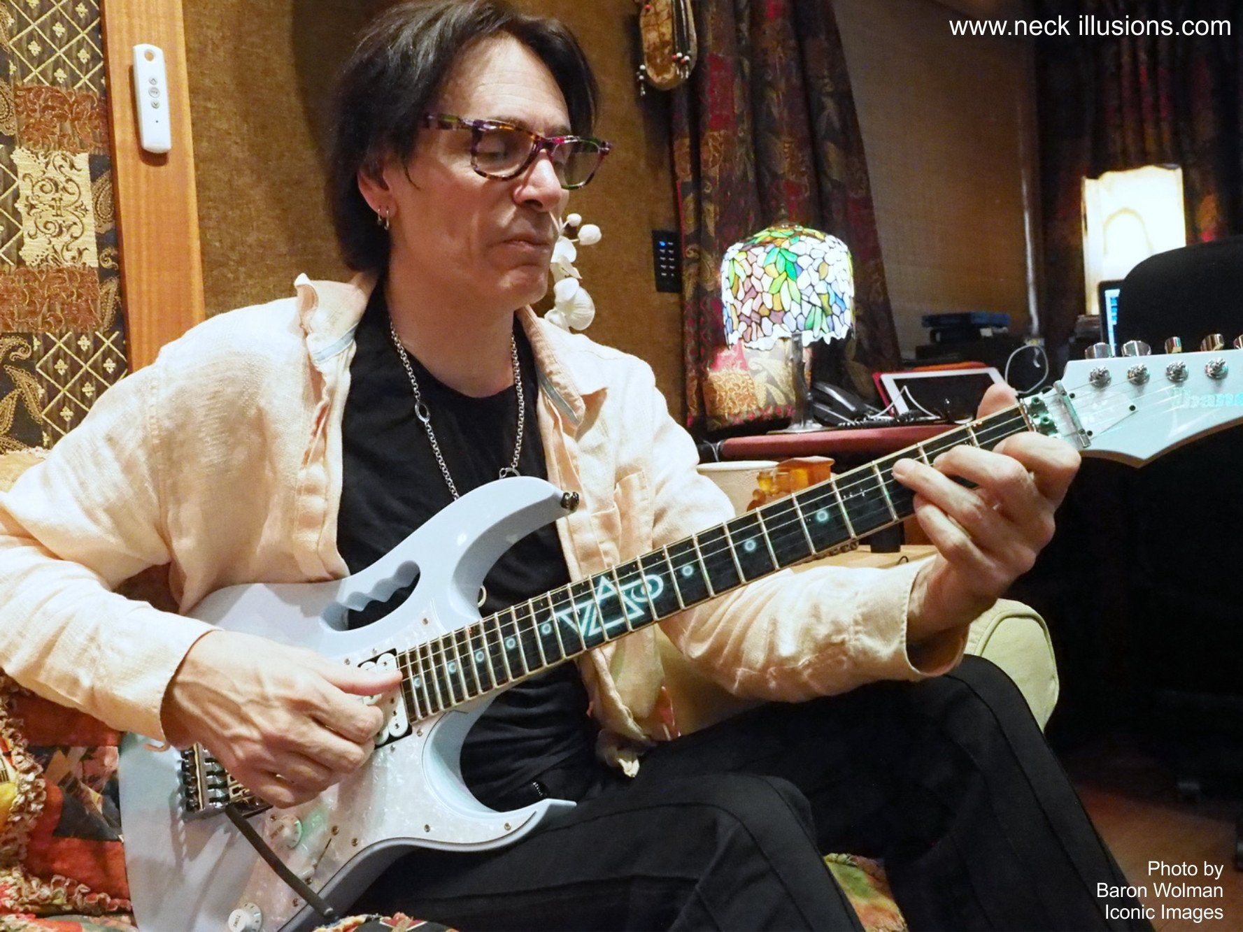 image of steve vai sitting in a recording studio playing a white electric guitar with a fret protector. the fret protector is black with dark colored symbols throghout. descending from the center of the fret protector is a glowing light blue and white steve vai logo. The steve vai logo makes the word "vai" with an upside down triangle, a right side up one, and a line going across the triangles with a curl at the end next to the triangle that is upright.