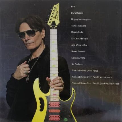 image of the back of the steve vai- modern primitive cd album artwork. there is an image of steve vai, wearing sunglasses and all black clothing. he holds a yellow electric guitar towards the camera. next to him in white text is the track listing for the album.