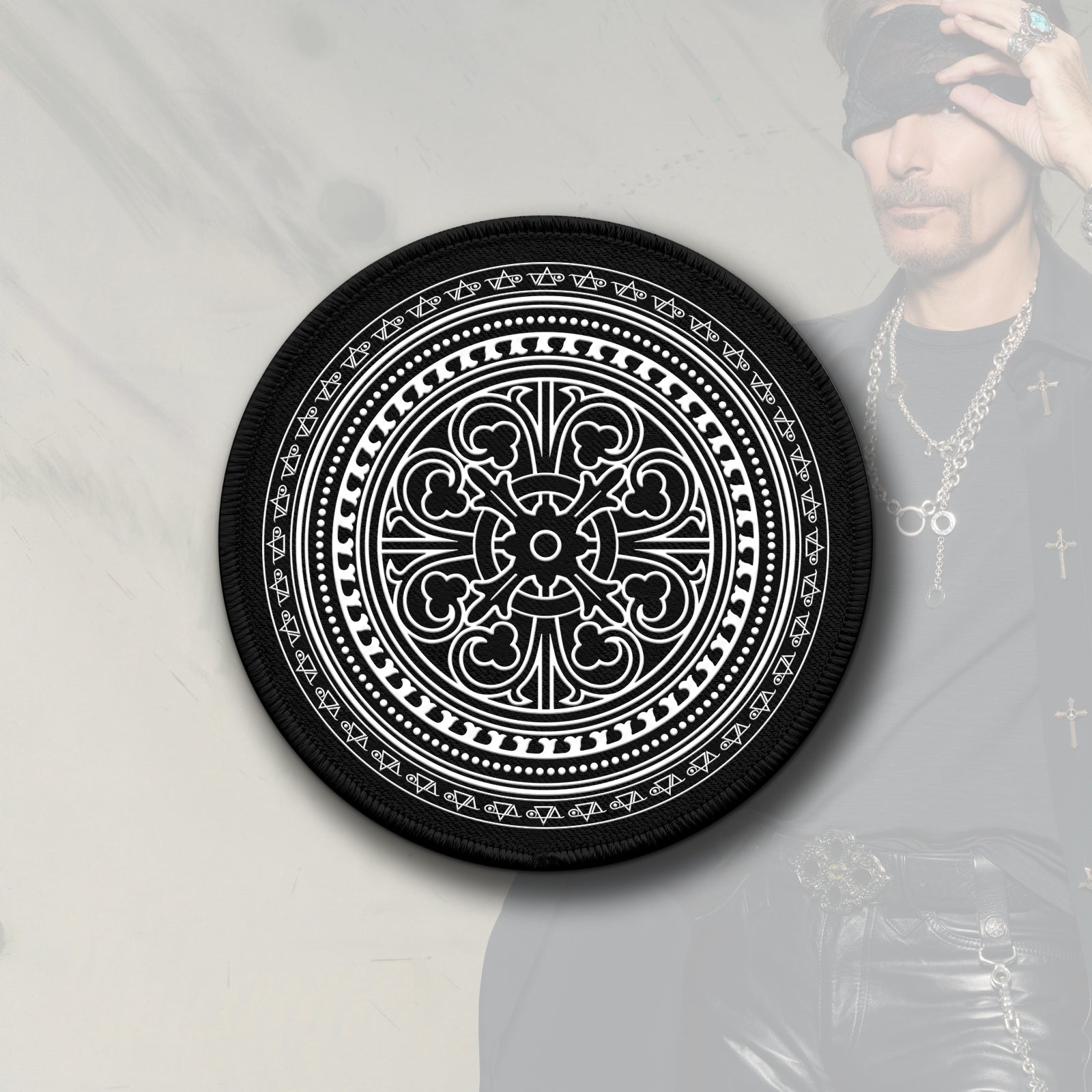  Image of a black and white circular patch against a faded white transparent background with an image of steve vai in the corner lifting a bandana up that is over his eye. the patch is black and has white print on it. it is an abstract circular image that has a pattern made up of various shapes and symbols that makes it look like it moves when you look at it.