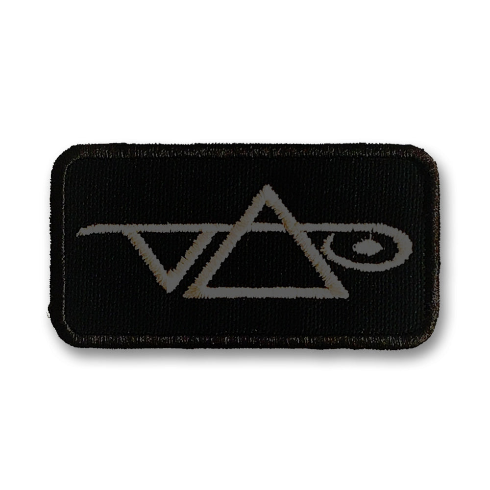 Image of a black patch against white background. the patch has a grey steve vai logo on it. The steve vai logo makes the word "vai" with an upside down triangle, a right side up one, and a line going across the triangles with a curl at the end next to the triangle that is upright.