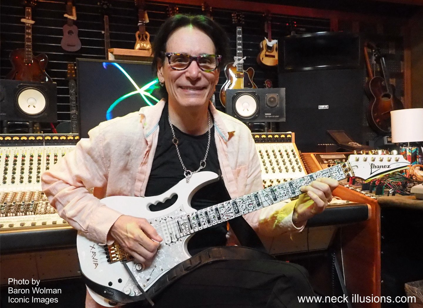 image of steve vai sitting on a chair in a recording studio looking at the camera and smiling. he holds a white electric guitar with a white fret protector on it that has pink/red, green, and black abstract symbols all over it.