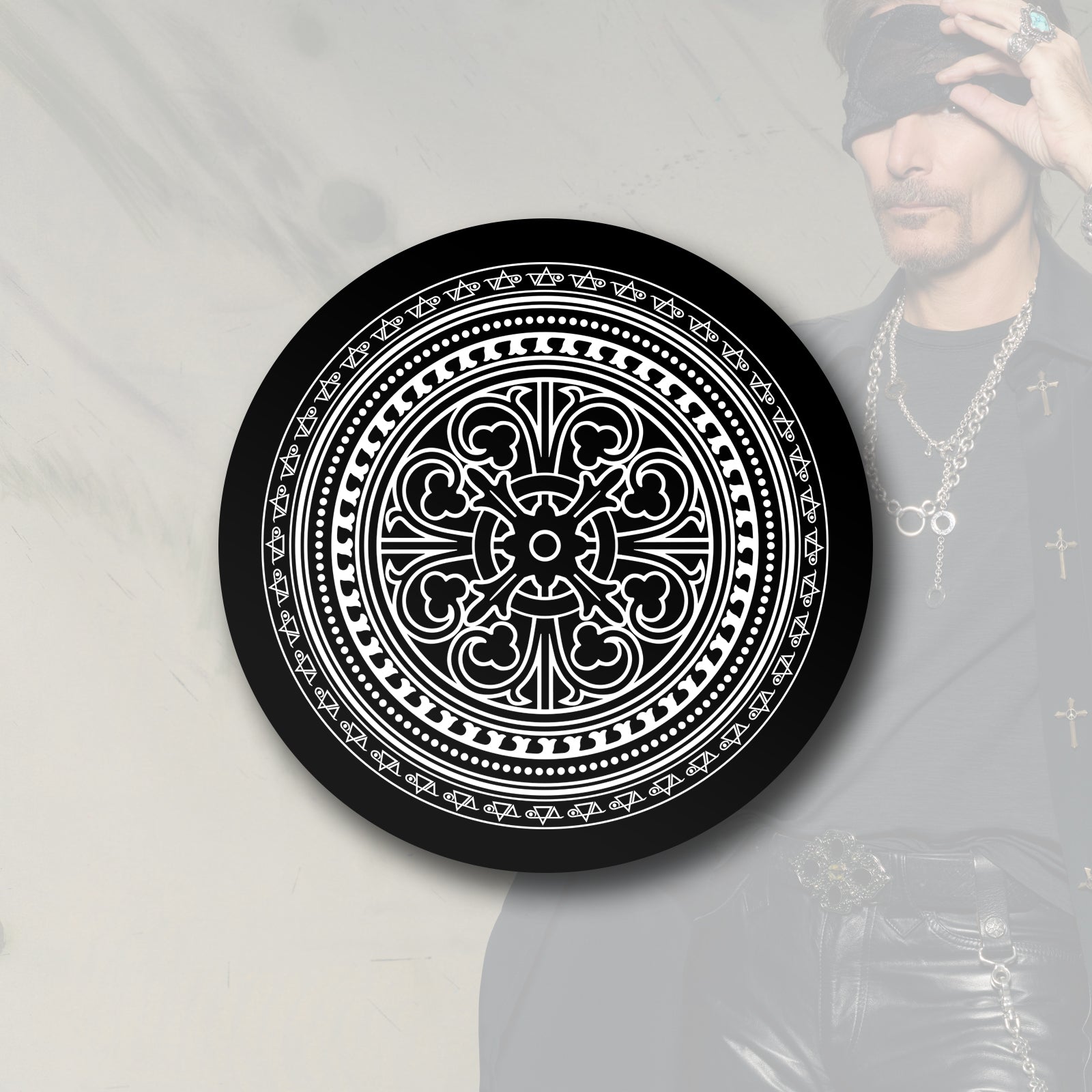 Image of a black and white circular sticker against a faded white transparent background with an image of steve vai in the corner lifting a bandana up that is over his eye. the sticker is black and has white print on it. it is an abstract circular image that has a pattern that makes it look like it moves when you look at it.