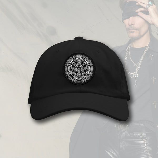 Image of a black dad hat with a black and white patch in the center against a faded white transparent background with an image of steve vai in the corner lifting a bandana up that is over his eye. the patch on the hat is black and has white print on it. it is an abstract circular image that has a pattern made up of various shapes and symbols that makes it look like it moves when you look at it.